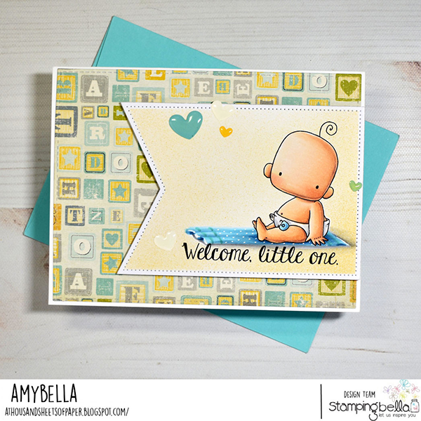 Handmade card design featuring Stamping Bella Baby Boy & Girl and Baby Sentiment Set cling mounted rubber stamp sets. The card shows a baby sitting on a blue polka-dotted blanket with the text 'Welcome, little one.' The background consists of a pattern of alphabet blocks in shades of green, yellow, and gray. The card is placed on top of a light blue envelope. Created by Amybella aka Amy Young from the blog 'A Thousand Sheets of Paper'.