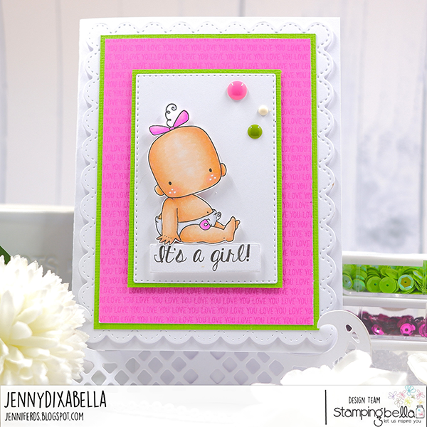 Handmade card design featuring a cute baby girl illustration from the Stamping Bella Baby Boy & Girl cling mounted rubber stamp set and a sentiment from the Baby Sentiment set. The card has a white scalloped border with a pink background that has the word 'LOVE' repeatedly printed in a subtle pattern. The central image is a baby girl with a pink bow on her head, sitting and wearing a diaper with a safety pin. The text 'It's a girl!' is written below the baby. The card is embellished with three enamel dots in pink, white, and green. Card designed by Jenny Dix aka JennyDixabella.