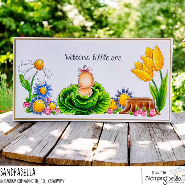 Handmade card design featuring the Stamping Bella Cabbage Patch Boy & Girl and Baby Sentiment Set cling mounted rubber stamp sets along with the Spring Backdrop rubber stamp. The card has a white background with colorful illustrations of flowers, a tree stump, and a baby sitting in a cabbage. The text on the card reads 'Welcome, little one.' The card is placed on a wooden surface with a blurred green outdoor background. The design is by Sandrabella.