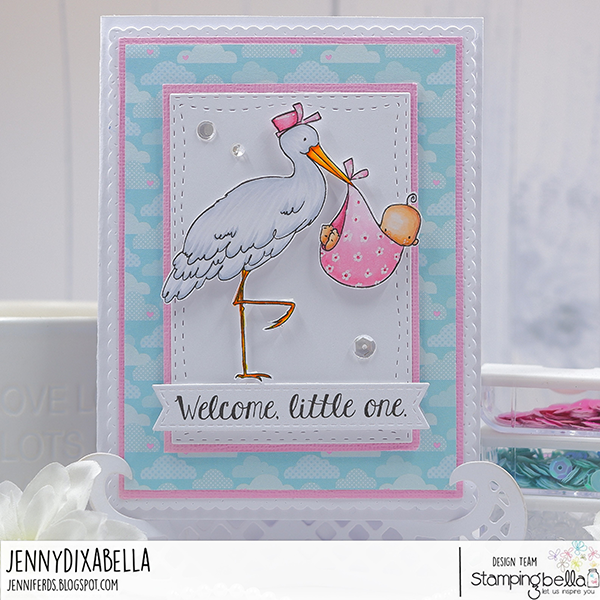 Handmade card design featuring Stamping Bella Delivery From the Stork cling mounted rubber stamp and the Baby Sentiment Set cling mounted rubber stamp set. The card showcases a stork wearing a pink hat, carrying a pink polka-dotted bundle with a baby inside. The background has a blue sky pattern with white clouds, and the card is bordered with a pink frame. The sentiment 'Welcome, little one.' is prominently displayed at the bottom. Card designed by Jenny Dixabella.