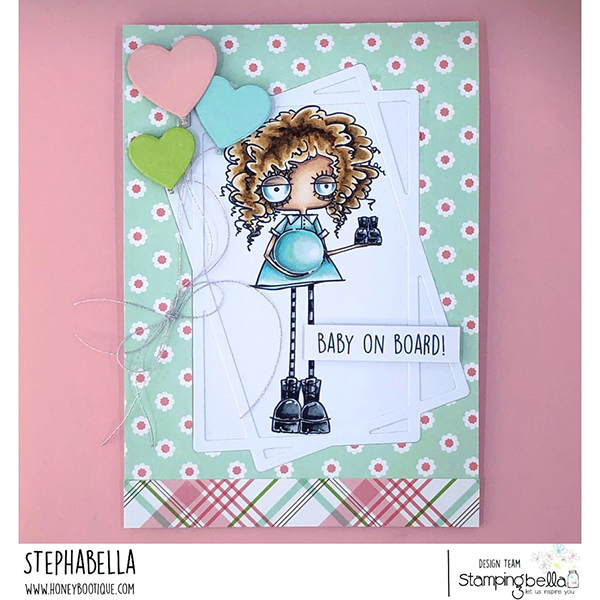 Handmade card design featuring Stamping Bella Oddball Baby on the Way cling mounted rubber stamp and Baby Sentiment Set cling mounted rubber stamp set. The card has a pastel green background with white and pink flowers, and a plaid pattern at the bottom. The central image is of a quirky, curly-haired character with a pregnant belly, wearing a blue dress and black boots. The character is holding a pair of baby shoes. Three heart-shaped balloons in pink, blue, and green are attached to the card with a string. The text 'BABY ON BOARD!' is prominently displayed on the card. The card is designed by Stephabella as part of the Stamping Bella design team.