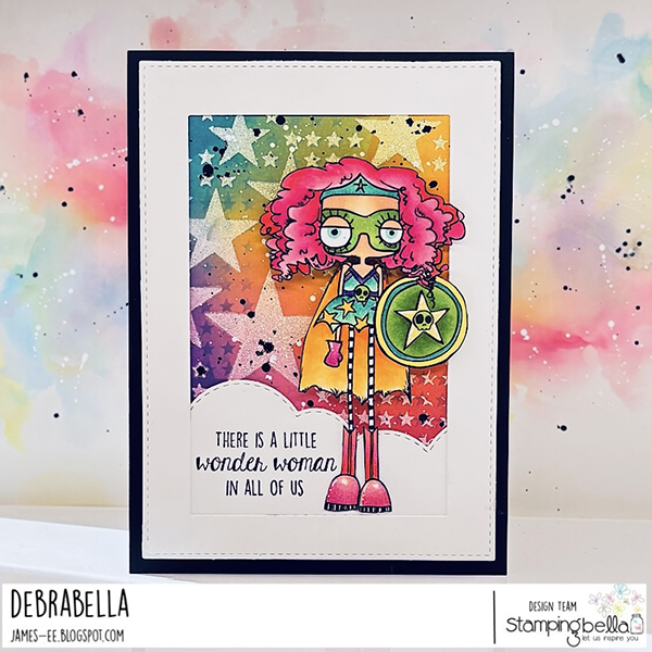 Handmade card design featuring the Stamping Bella Oddball Girl Power cling mounted rubber stamp. The card has a rainbow gradient background with star patterns. A quirky girl with pink curly hair, green mask, star headband, yellow cape, and pink boots stands at the center, holding a green shield with a star emblem. Sentiment below reads 'There is a little wonder woman in all of us.' Framed with a white border and black background. Created by Debrabella from Stamping Bella design team.