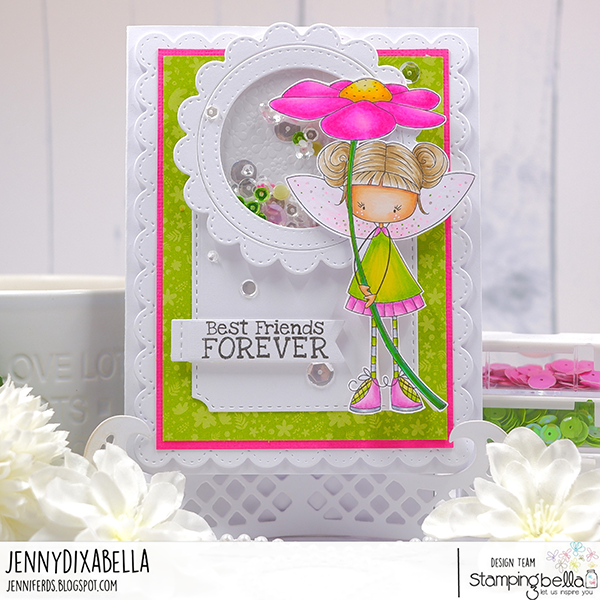 Handmade shaker card design featuring Stamping Bella Whimsabella DaisyDream and Spec-Tacular Together Sentiment Set cling mounted rubber stamps. The card has a white scalloped border with a green background and pink accents. A fairy character with blonde hair in buns, wearing a green and pink outfit, holds a large pink flower. The sentiment 'Best Friends FOREVER' is stamped on a white tag. The card is embellished with sequins and beads, adding a playful and whimsical touch. The design is showcased on a white decorative stand with flowers in the foreground. Design by Jenny Dix for Stamping Bella.