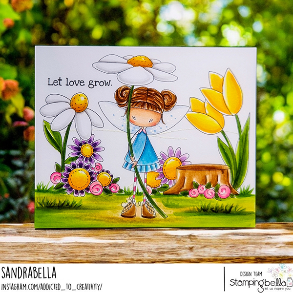 Handmade card design featuring Stamping Bella Whimsabella DaisyDream, Spring Backdrop, and Garden Sentiment Set cling mounted rubber stamps. The card showcases a whimsical fairy girl in a blue dress holding a large daisy, surrounded by colorful flowers, a tree stump, and lush greenery. The background is a vibrant garden scene with large white daisies, yellow tulips, and purple flowers. The sentiment 'Let love grow.' is stamped in the top left corner. The card is designed by Sandrabella, as part of the Stamping Bella design team.