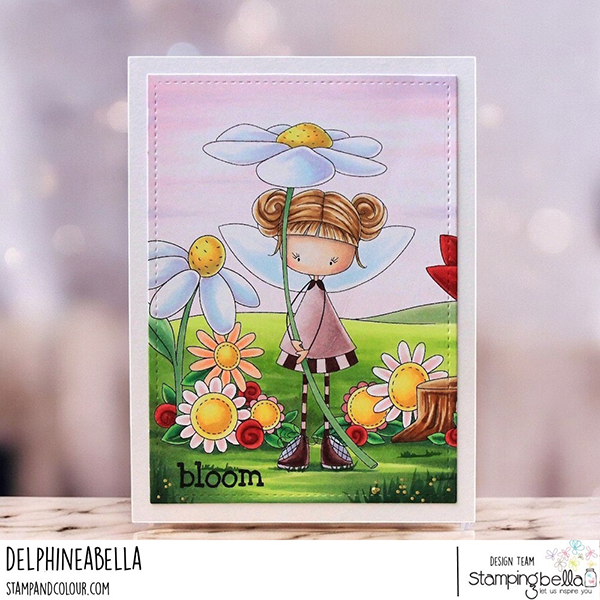 Handmade card design featuring Stamping Bella Whimsabella DaisyDream, Spring Backdrop, and Hello Spring Sentiment Set cling mounted rubber stamps. The card showcases a whimsical fairy girl with blonde hair in buns, wearing a pink dress and striped stockings, holding a large daisy flower. She is surrounded by a vibrant spring scene with various daisies, green grass, and a tree stump. The word 'bloom' is stamped at the bottom left corner. The background includes a soft pink sky and rolling green hills, creating a cheerful and enchanting springtime atmosphere. The card is designed by Delphineabella as part of the Stamping Bella design team.