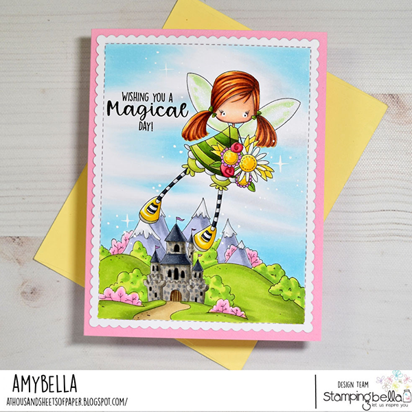 Handmade greeting card featuring Stamping Bella Whimsabella GlimmerGlade, Fairytale Backdrop, and Fairytale Sentiment Set cling mounted rubber stamps. The card shows a whimsical fairy girl with red hair holding a bouquet of flowers, flying over a fairytale landscape with a castle, green hills, and mountains. The scene is colored with Copic alcohol markers in vibrant hues. The sentiment 'Wishing you a magical day!' is stamped on the upper left side. The card is bordered with a pink scalloped edge and placed on top of a yellow envelope. Designed by Amybella from the blog 'A Thousand Sheets of Paper.' for the Stamping Bella design team.