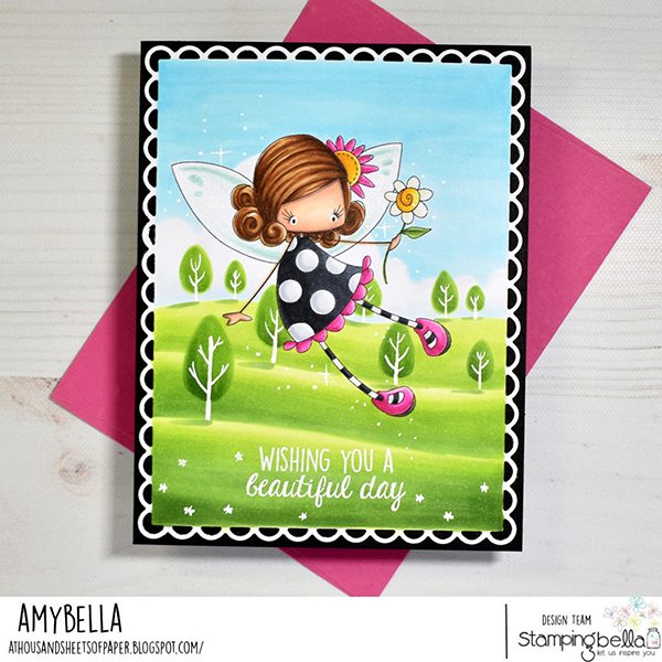 Handmade greeting card featuring a whimsical fairy scene colored with Copic alcohol markers. The card showcases the Stamping Bella Whimsabella PetalShimmer cling mounted rubber stamp and Hello Spring Sentiment Set cling mounted rubber stamp set. The fairy has brown hair styled in curls, wears a black dress with white polka dots, and has pink shoes. She is holding a yellow flower and has translucent wings with sparkles. The background is a bright, cheerful landscape with green hills and stylized trees under a blue sky. The card has a scalloped white border on a black background and includes the sentiment 'Wishing you a beautiful day' at the bottom. The card is displayed on a pink envelope. The design is by Amybella from the Stamping Bella design team.