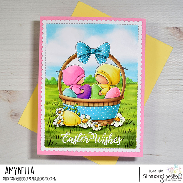 Handmade Easter greeting card featuring Stamping Bella Bundle Girls in the Easter Basket cling mounted rubber stamp. The card shows two adorable characters dressed in pink and yellow outfits sitting inside a blue polka-dotted Easter basket with colorful eggs. The basket is adorned with a large blue bow and surrounded by daisies and greenery. The background depicts a bright, grassy field under a clear blue sky. The card has a pink border and the text 'Easter Wishes' is written at the bottom in elegant white script. Created by Amy Young for the Stamping Bella design team.