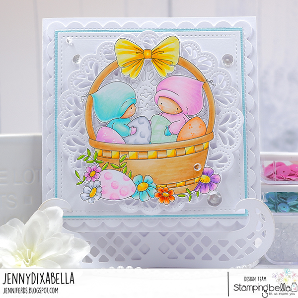 Handmade greeting card featuring Stamping Bella Bundle Girls in the Easter Basket cling mounted rubber stamp. Two cute girls in pastel hooded outfits sit in a wicker basket with a yellow bow, surrounded by decorated Easter eggs and vibrant spring flowers. The card has a white lace-like border and is embellished with sequins for added sparkle, set against a white background with a blue border. Created by Jenny Dix for the Stamping Bella design team.