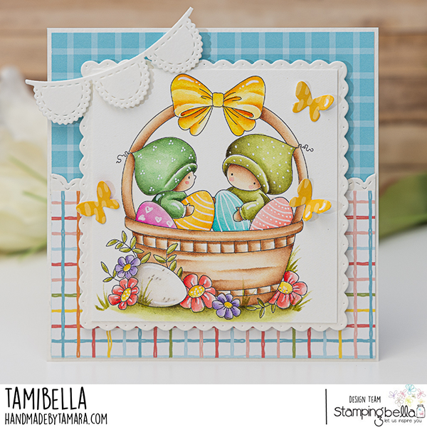 Handmade Easter greeting card featuring Stamping Bella Bundle Girls in the Easter Basket cling mounted rubber stamp. The card showcases two adorable girls in green hats sitting in a wicker basket filled with colorful Easter eggs. The basket is adorned with a large yellow bow and surrounded by vibrant flowers and butterflies. The background includes a blue checkered pattern and a multicolored plaid design, with a decorative white scalloped edge and banner. The card is designed by Tamara for Stamping Bella.