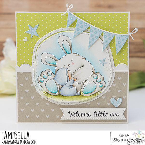 Handmade greeting card featuring a cute illustration of a bunny and a small child cuddling, created using Stamping Bella Bundle Girl loves Bunny Wobbles cling mounted rubber stamp. The card has a green polka dot background with blue star and heart embellishments, and a banner of blue polka dot flags. The bottom section has a beige background with white hearts and a banner that reads 'Welcome, little one.' Card designed by Tamibella for the Stamping Bella design team.