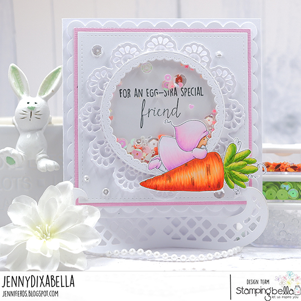 Handmade greeting card featuring Stamping Bella Bundle Girl with a Carrot cling mounted rubber stamp. The card showcases a cute illustration of a girl in a pink hooded outfit hugging a large orange carrot with green leaves. The card is decorated with a white doily-like circular frame, sequins, and the sentiment 'For an egg-stra special friend.' The background includes a white ceramic bunny and a white flower, adding to the Easter theme. The card was designed by Jenny Dixabella for Stamping Bella.